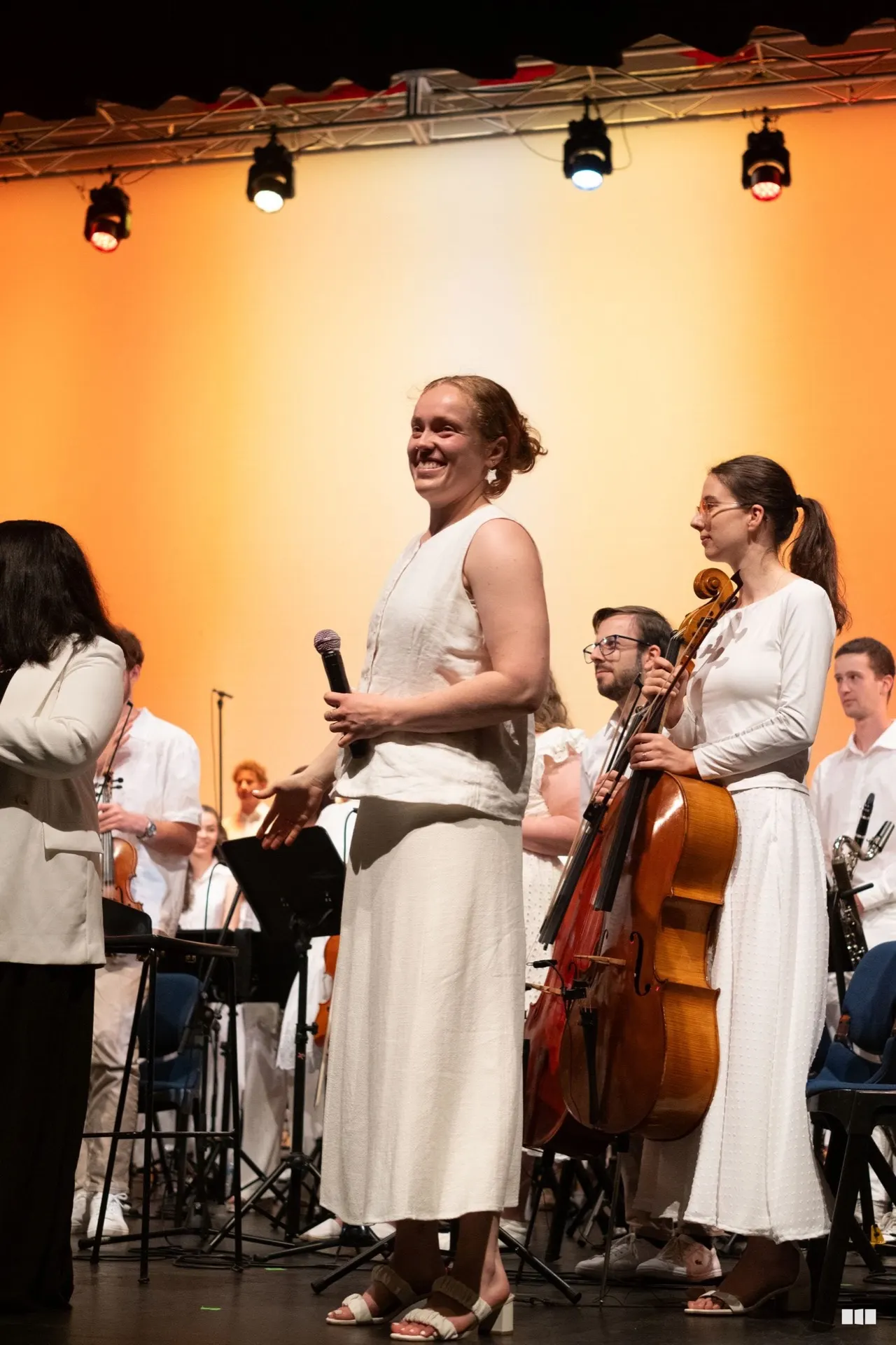 A presenter addressing the audience mid-concert with the orchestra behind her