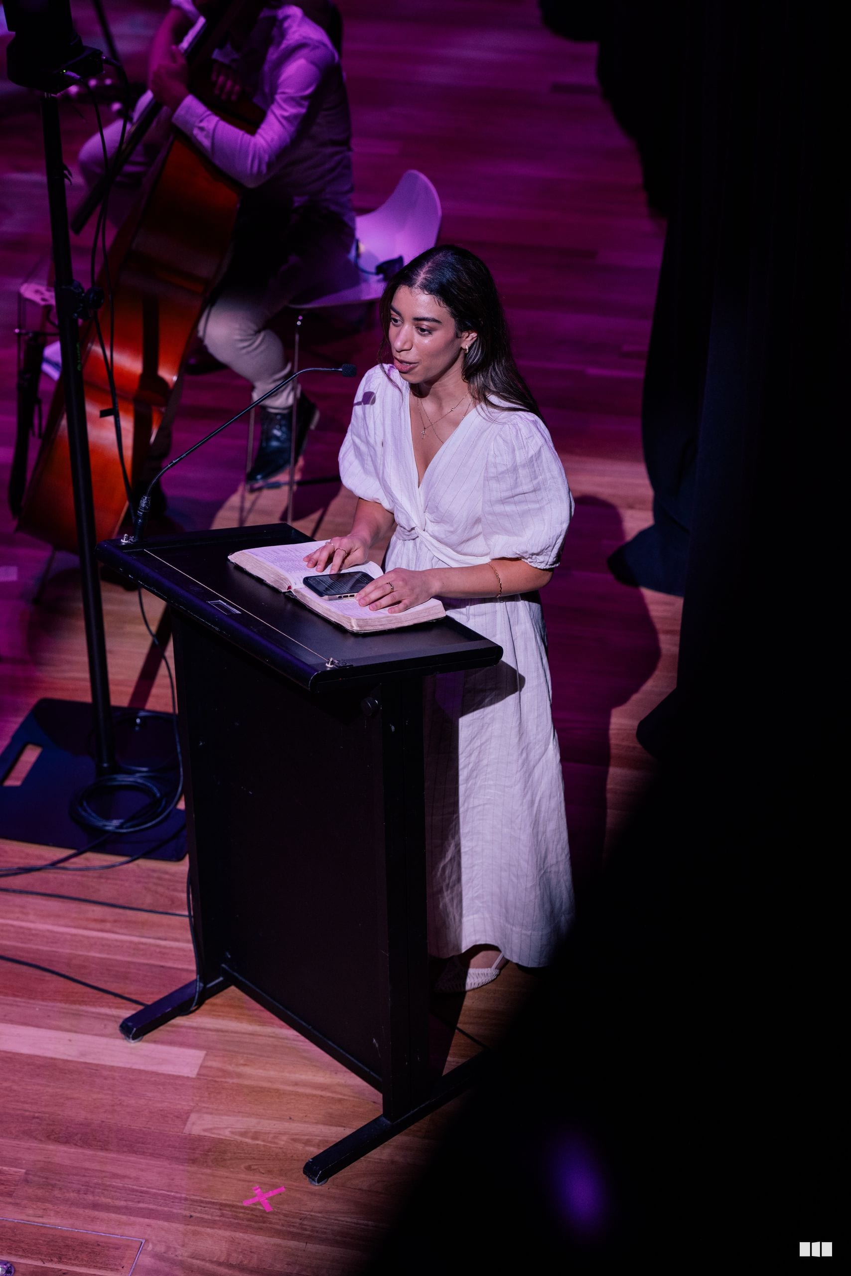 A woman in a white dress reads from a document at a podium during a performance with orchestra musicians and purple stage lighting in the background.