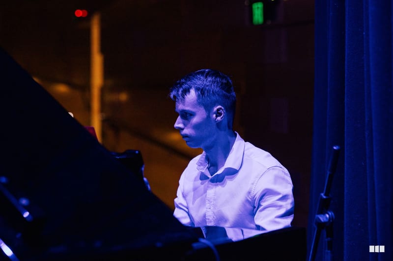 A pianist in a white shirt performs at the keyboard during an evening concert, illuminated by blue stage lighting.