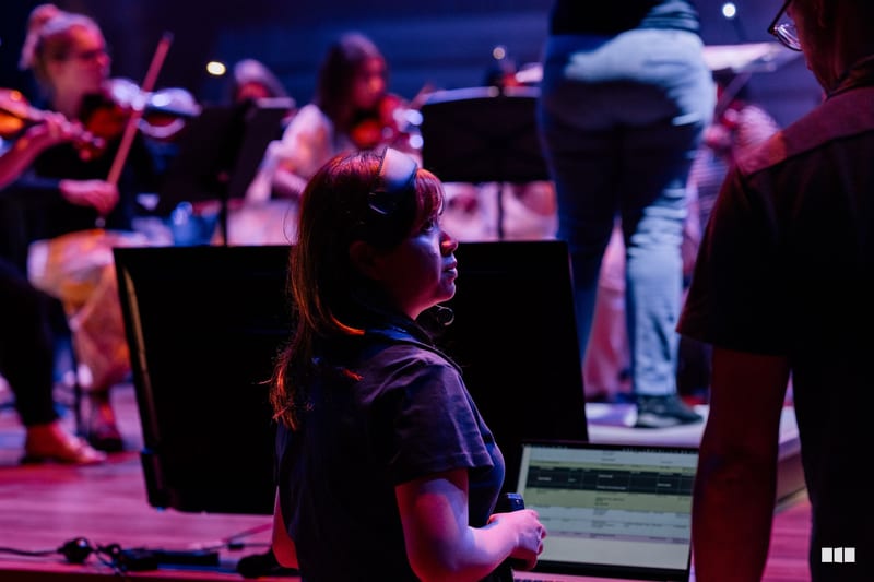 A young performer stands engaged during an orchestral rehearsal or performance with musicians and instrumental equipment visible in the atmospheric stage lighting.