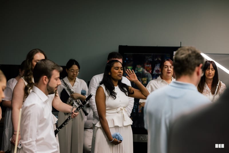A choir member in white attire gestures expressively while rehearsing with ensemble members in a studio setting.