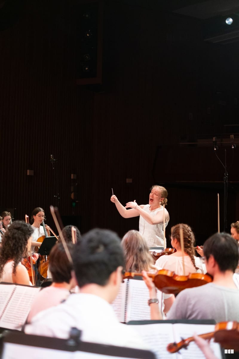 A conductor leads a string orchestra during rehearsal in a concert hall with sheet music visible on stands.
