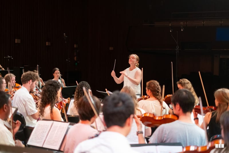 A conductor leads a string orchestra during rehearsal, with musicians holding violins and violas while reading from sheet music.
