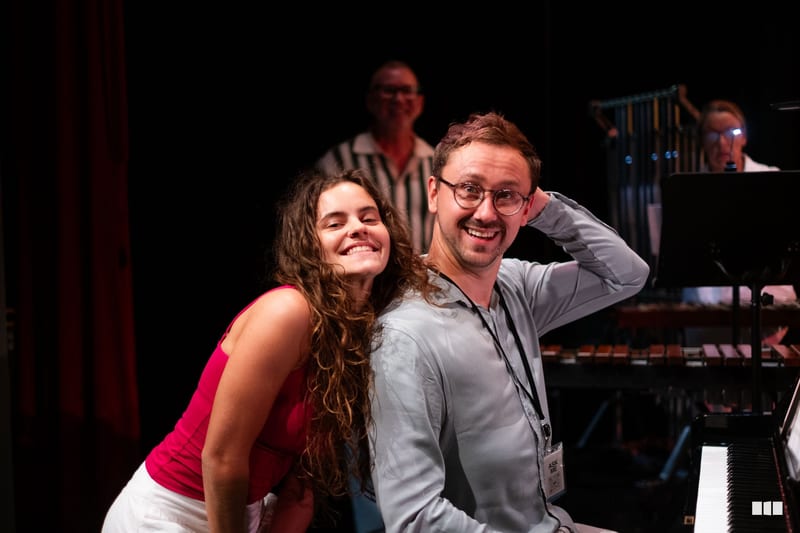 A young woman in a pink top and a man wearing glasses and a lanyard smile together on stage near a piano, with other musicians visible in the background.