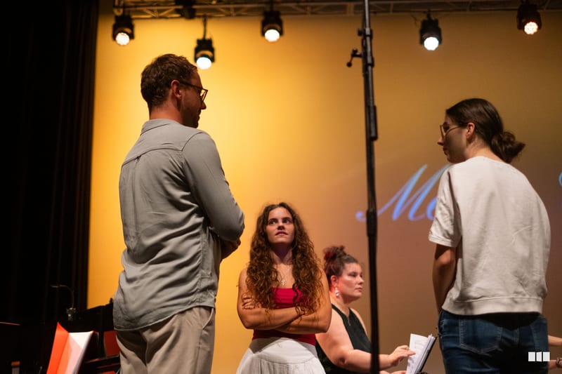 A group of young performers engage in discussion during a rehearsal or workshop on stage under warm lighting.