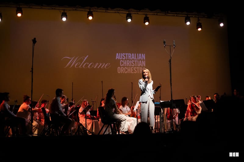 A female soloist in white performs at the microphone with the Australian Christian Orchestra accompanying her on stage.