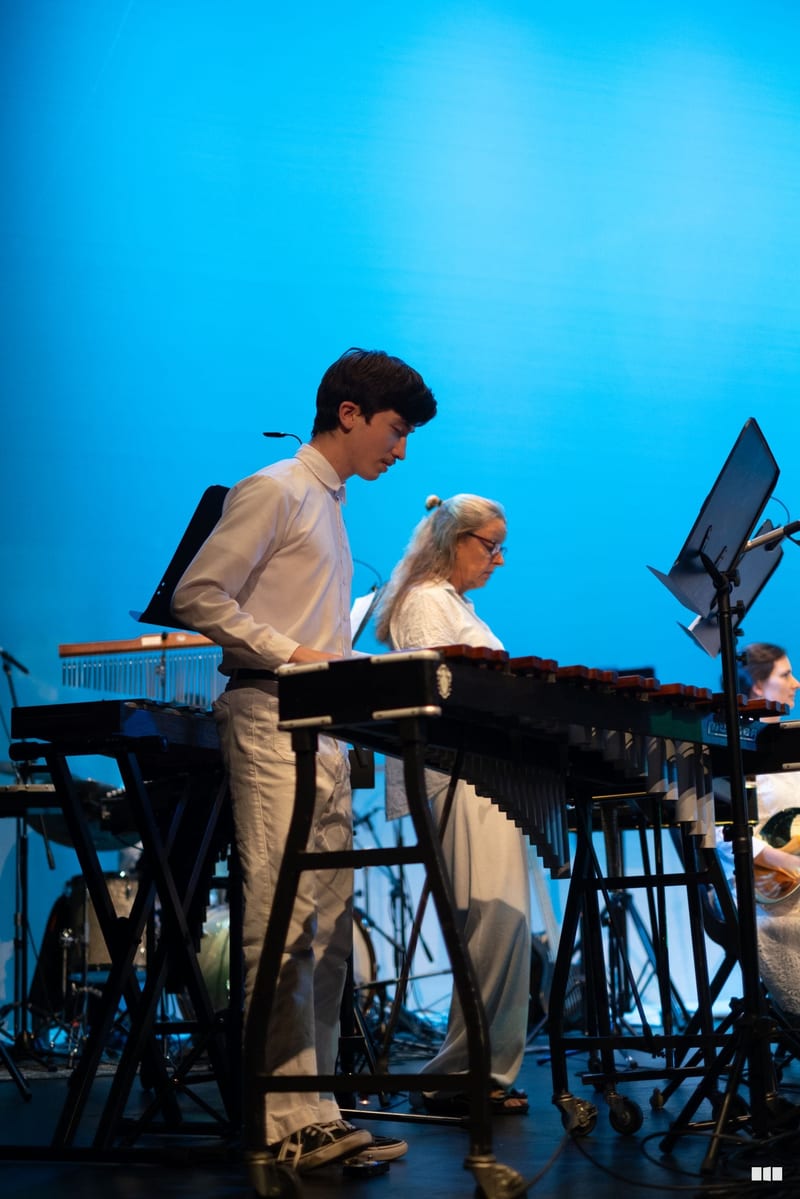 Young percussionist performs at a vibraphone alongside an older musician on stage against a bright blue backdrop.