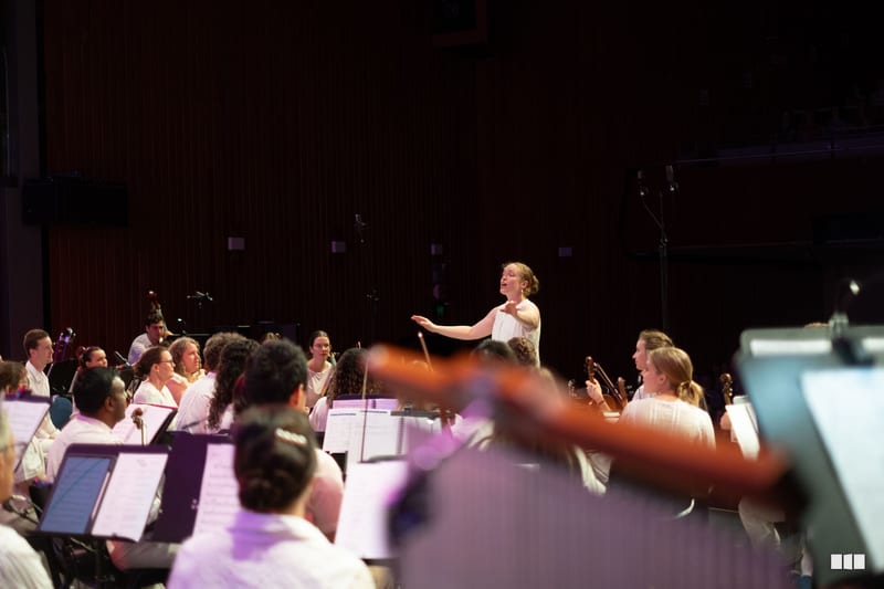 A conductor leads the orchestra during a performance, with musicians holding sheet music and instruments in a dimly lit concert hall.