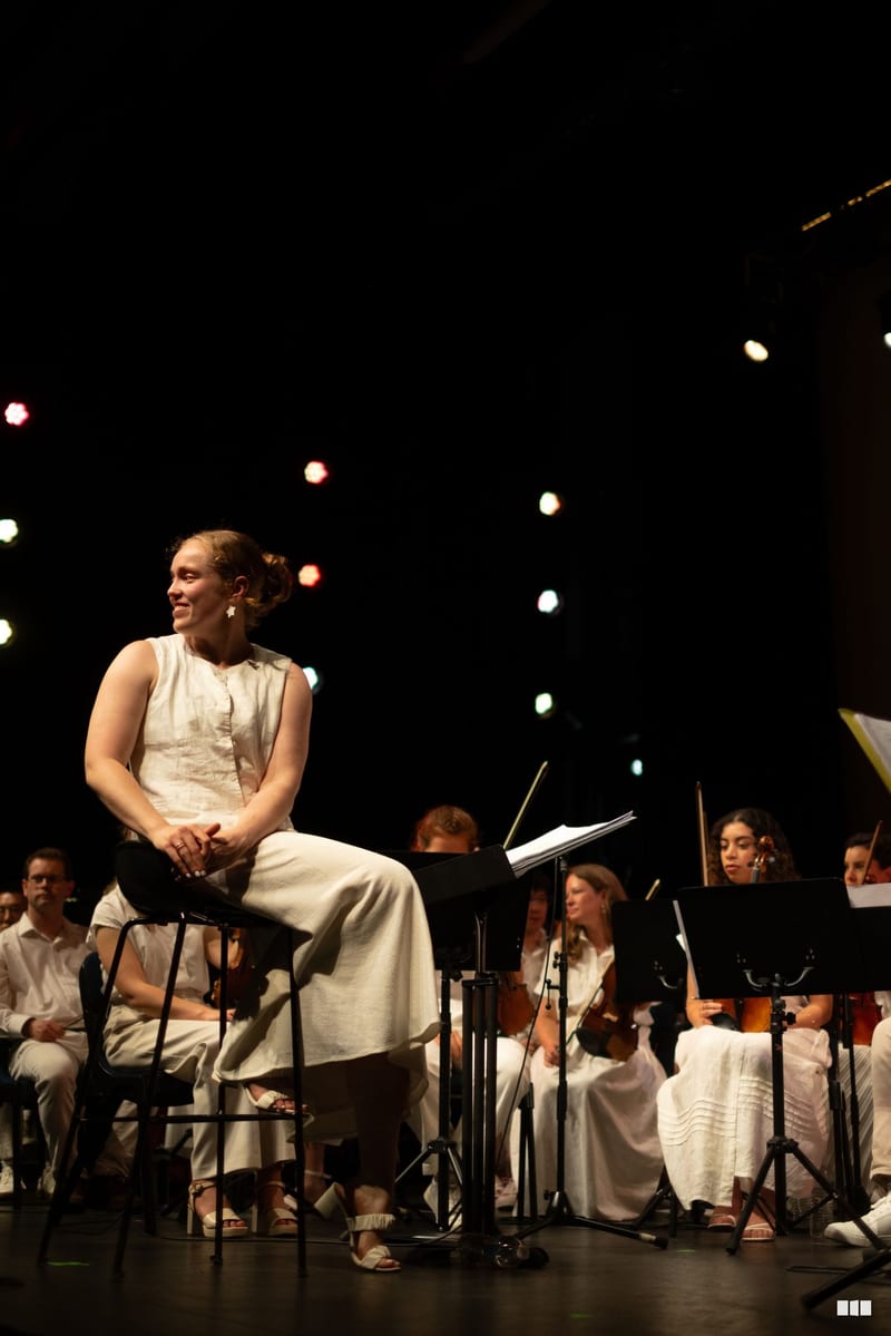 A female soloist sits on stage surrounded by orchestra members during a performance against a backdrop of warm stage lighting.