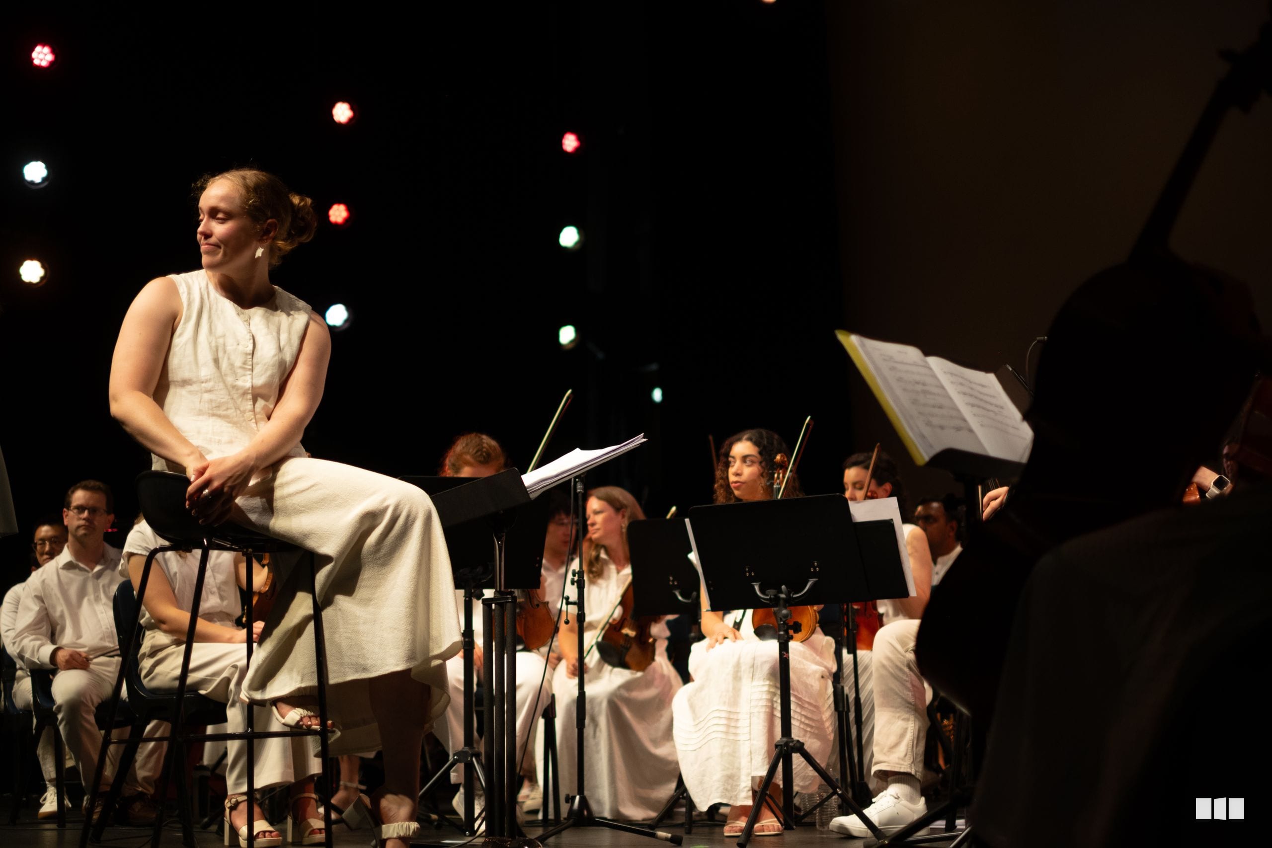 A soloist in a cream-colored dress performs with an orchestra during a concert, with atmospheric stage lighting in the background.