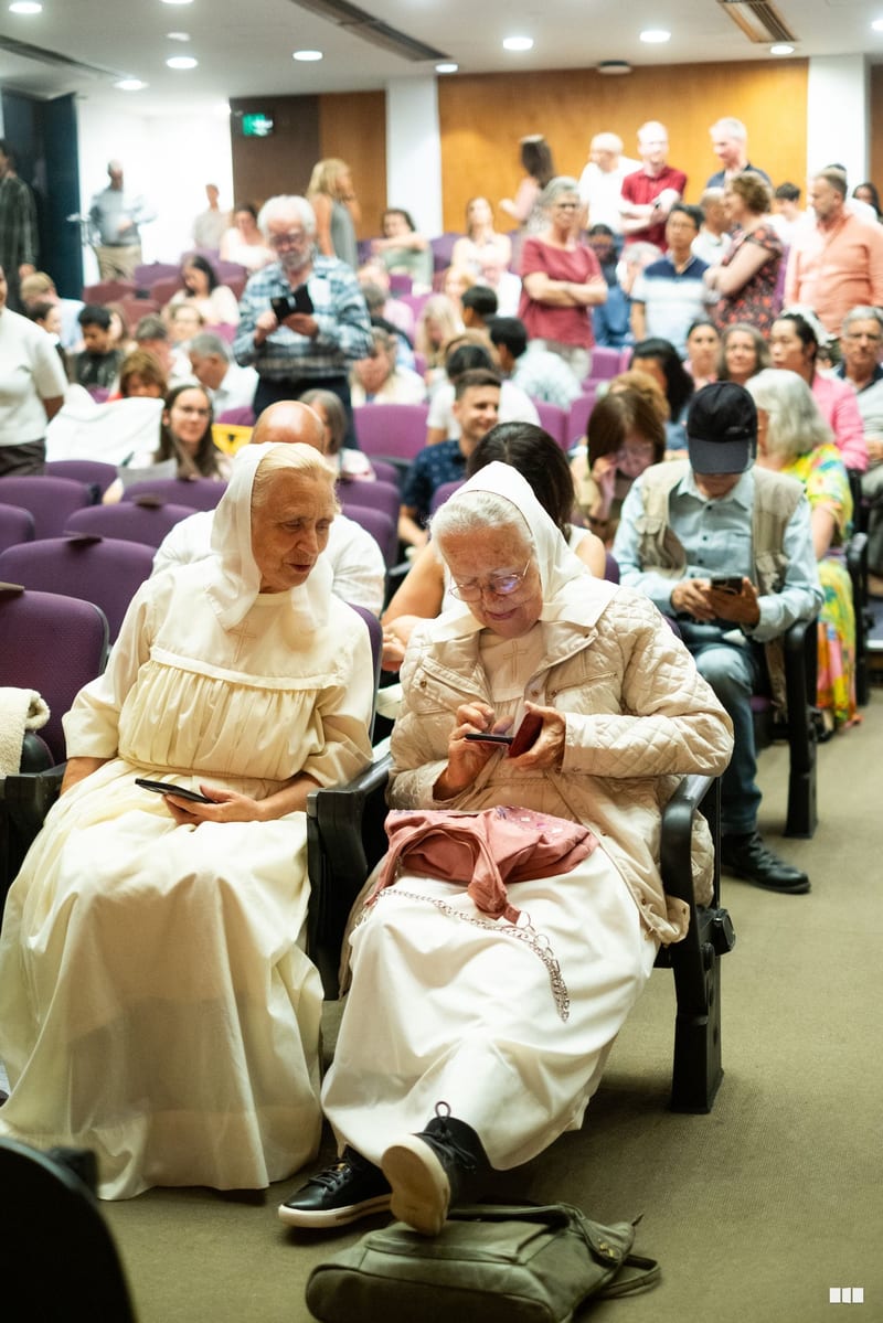 Two elderly women in traditional religious attire sit together during a community gathering in an auditorium filled with attendees.