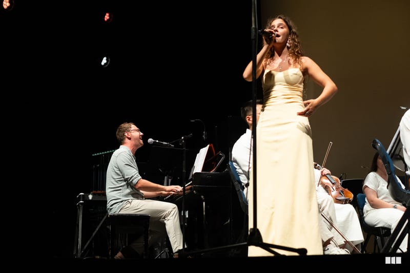 A female soloist in a cream gown performs with a pianist and string musicians on stage.