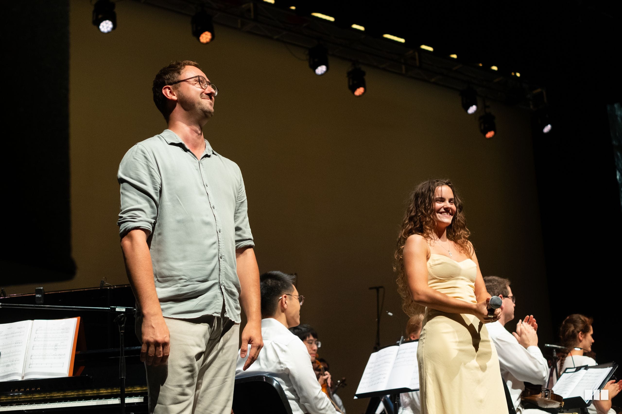 A male soloist in a light blue shirt stands beside a female soloist in a cream dress holding a microphone, with orchestra members and sheet music visible on stage.