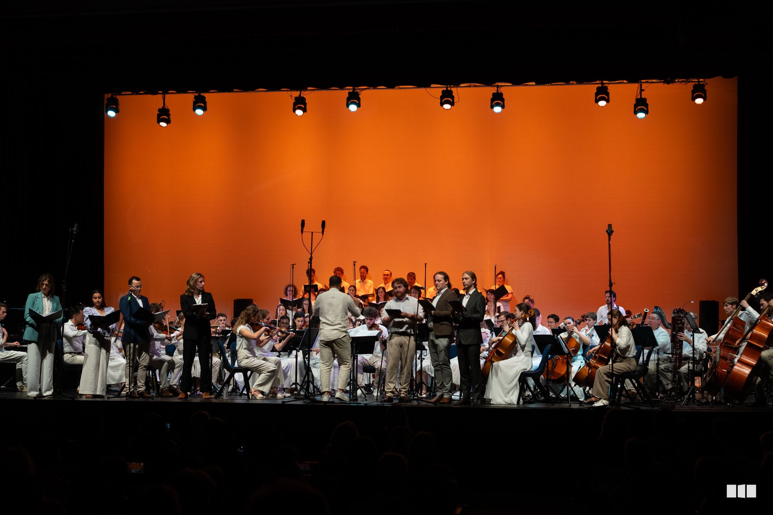 A conductor leads a large orchestra and choir during a performance on stage with an orange backdrop and professional lighting.