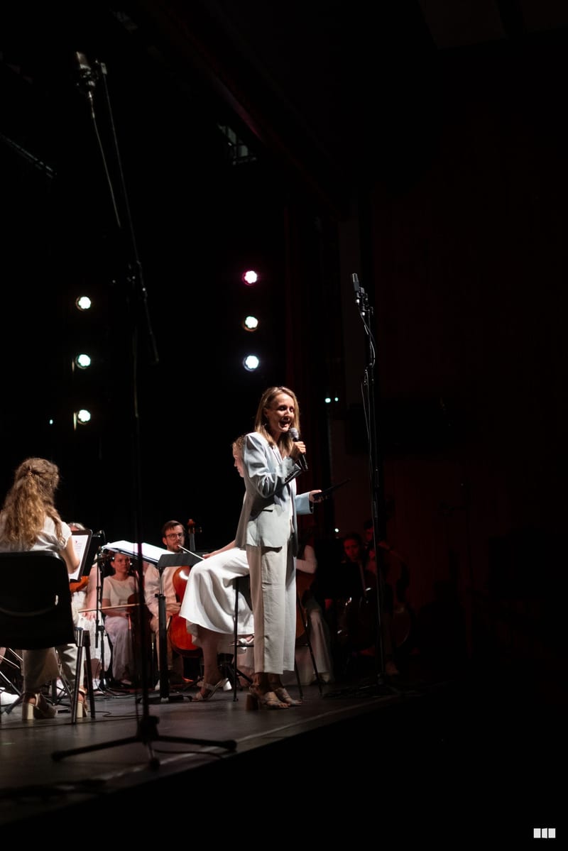 A soloist in a light blue dress performs at the microphone with orchestra members seated beside her on stage under dramatic theatrical lighting.