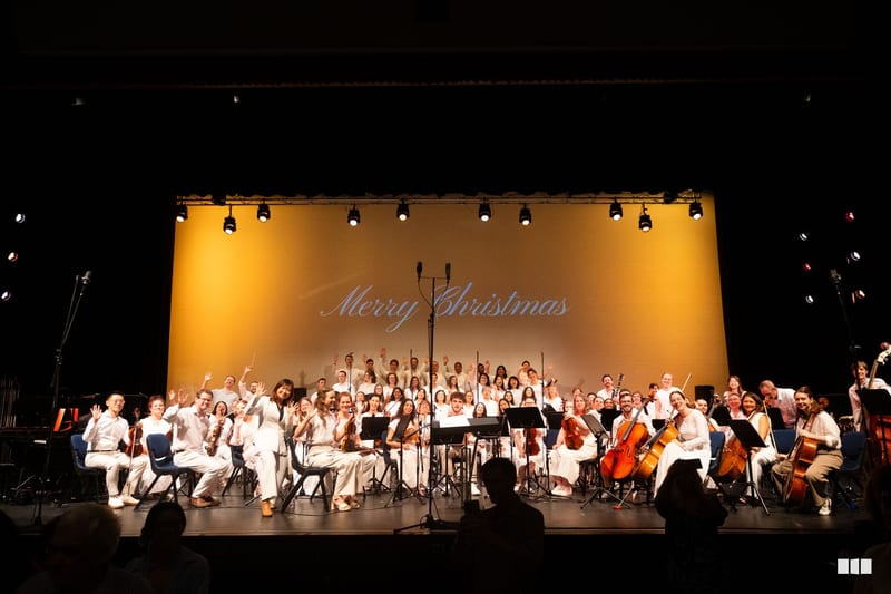 A large orchestra and choir perform a festive Christmas concert on stage under warm golden lighting with 'Merry Christmas' displayed on the backdrop.