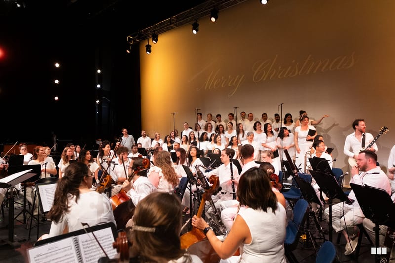 A large orchestra and choir in white attire perform together on stage during a Christmas concert, with musicians arranged in traditional orchestral formation.