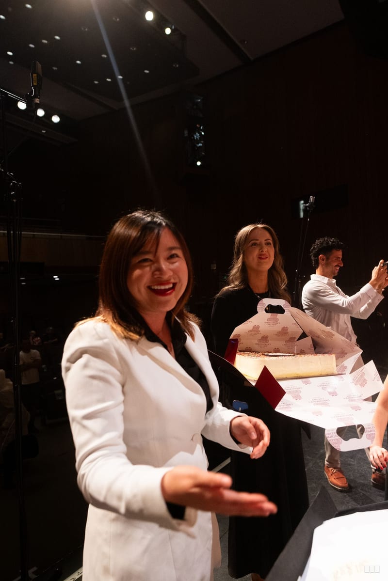 A woman in a white blazer smiles at the camera during what appears to be a formal event or rehearsal in a concert venue.