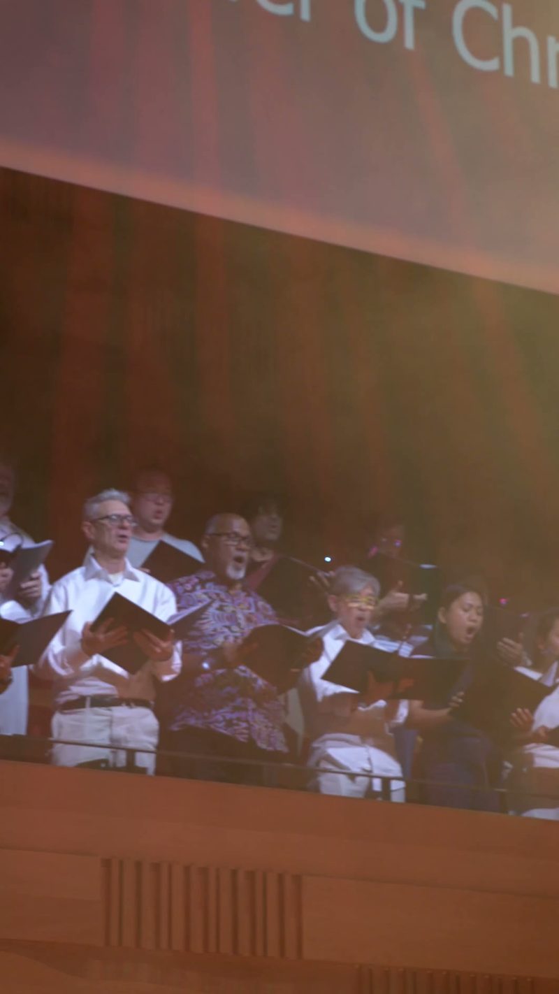 Choir members holding hymnals and singing together during a performance in front of a projected screen.