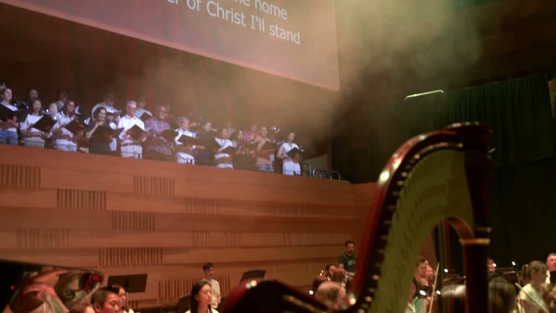 A choir sings in the upper balcony while orchestra members perform in the foreground during a worship service, with a projected hymn lyric displayed above.