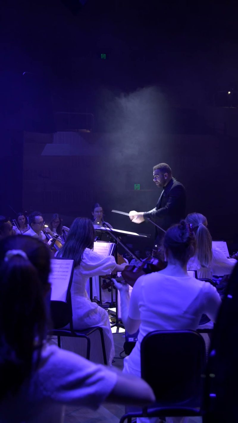 A conductor leads an orchestra in a dramatic blue-lit performance with musicians seated and reading from sheet music.