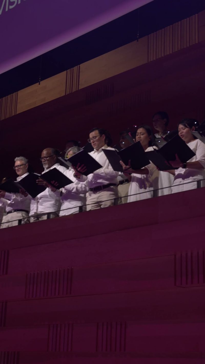 A choir performs on stage in formal attire, holding sheet music and singing together under atmospheric purple lighting.