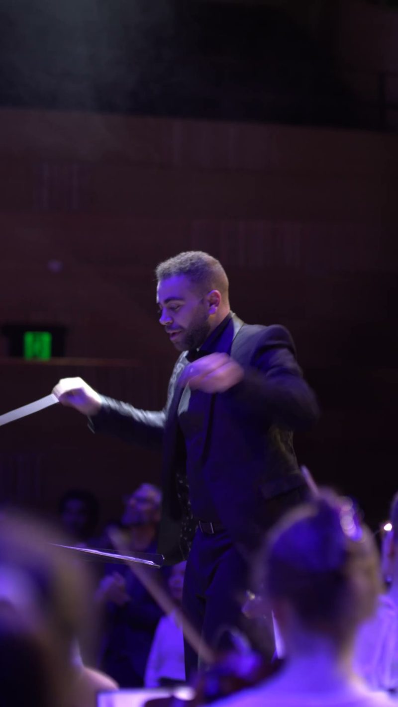 A conductor in formal attire leads the orchestra with an animated gesture during a performance under purple stage lighting.