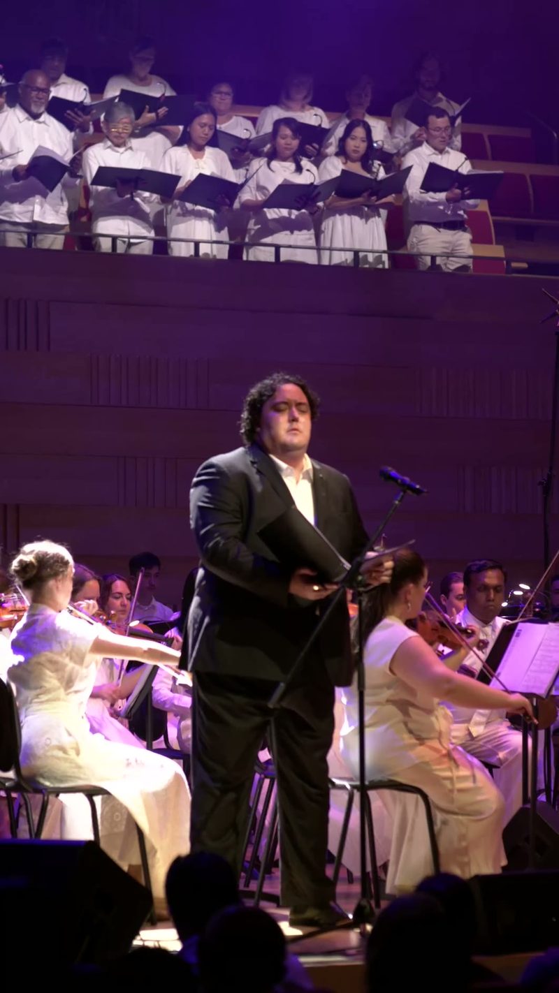 A conductor leads the orchestra and choir in performance, with singers in white attire positioned above and ensemble musicians below on stage.