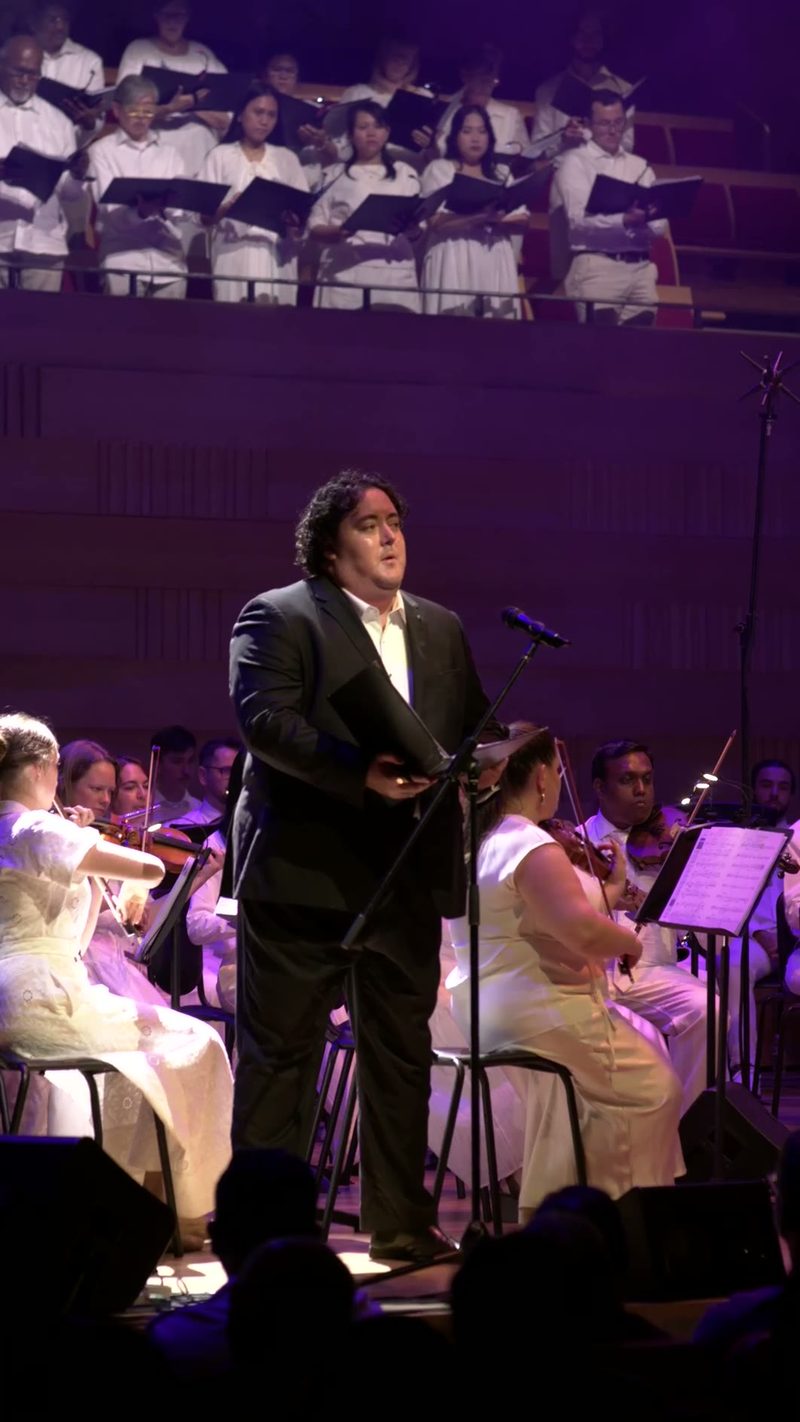 A male soloist in formal attire performs at the microphone accompanied by an orchestra and choir in a concert hall setting.