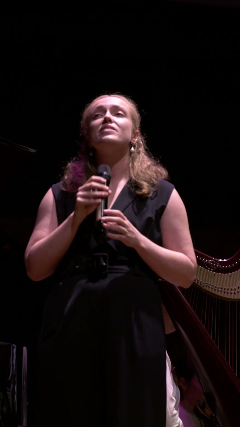 A female soloist holds a microphone while performing on stage with a harp visible in the background.