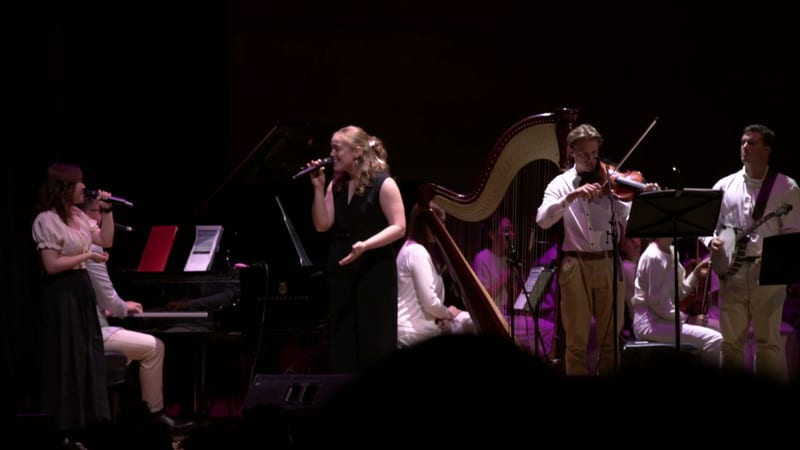 A chamber ensemble performs with piano, harp, violin, and vocalists on stage under purple lighting.