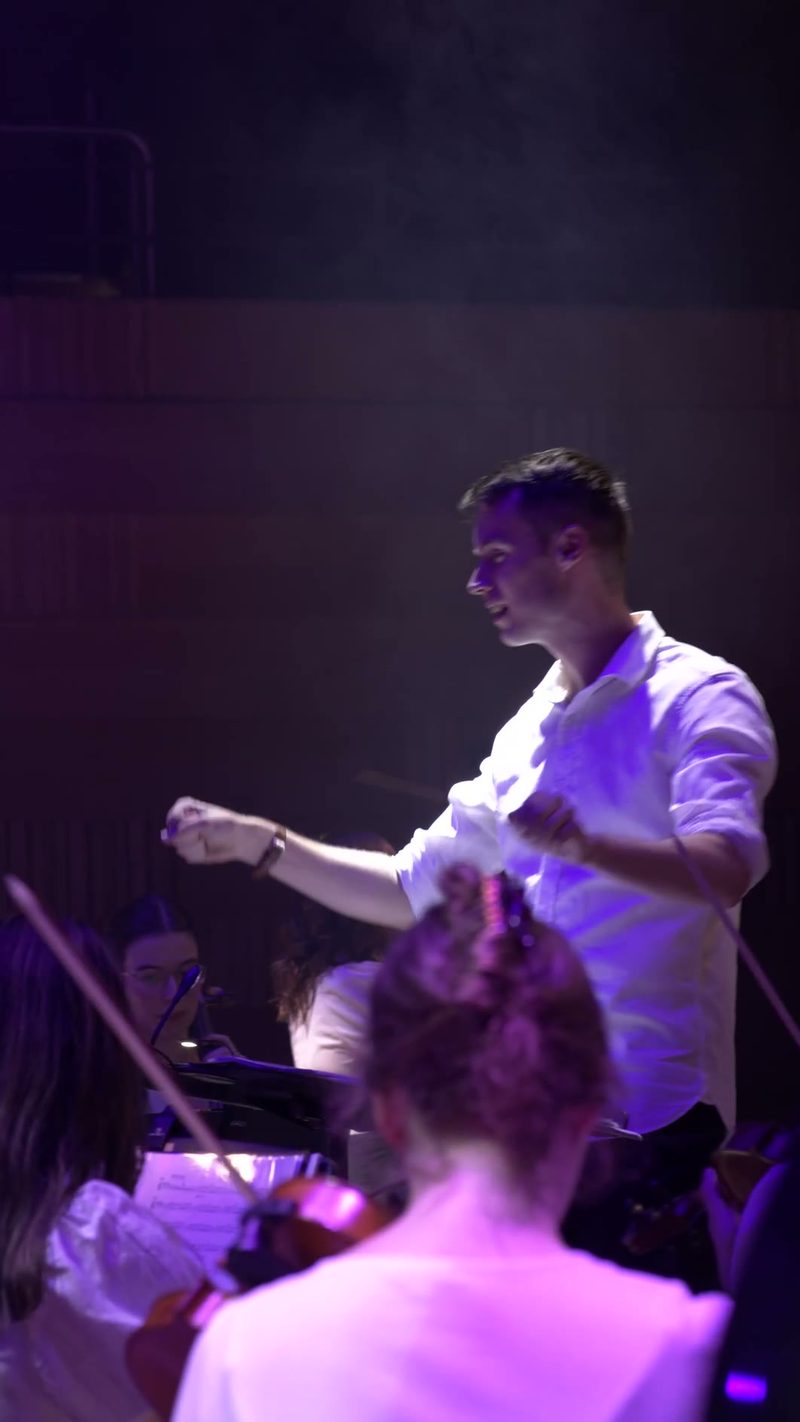 A conductor leads an orchestra during a performance with purple stage lighting, engaging with musicians in the ensemble.