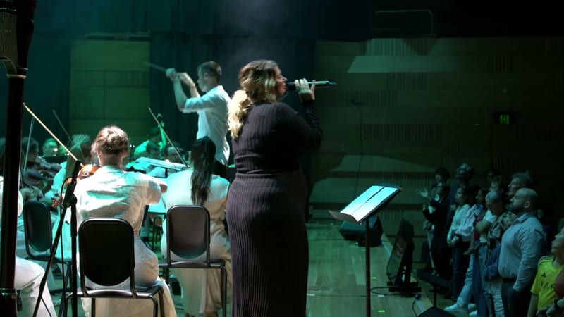 A female soloist in a dark pinstriped outfit performs at the microphone while ensemble members accompany her on stage.