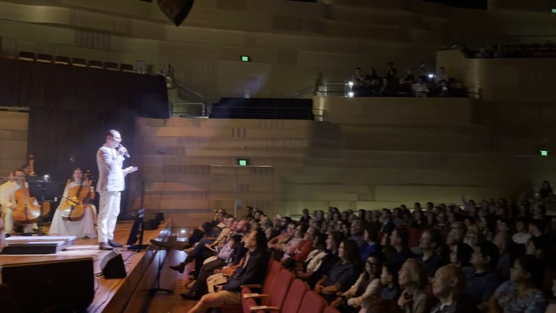 A conductor in a white jacket addresses the audience during a classical music performance in a concert hall.