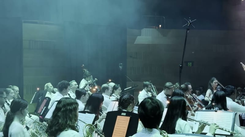 A large orchestra with string, woodwind, and brass sections performs together in a concert setting with atmospheric stage lighting.