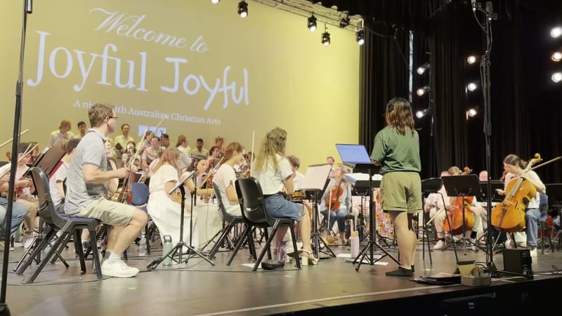 Young orchestra musicians perform at a Joyful Joyful concert presented by South Australian Christian Arts, with string and wind players seated on stage under professional lighting.