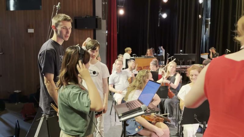 Young musicians and conductor engage with an audience member holding a laptop during an interactive orchestra presentation.