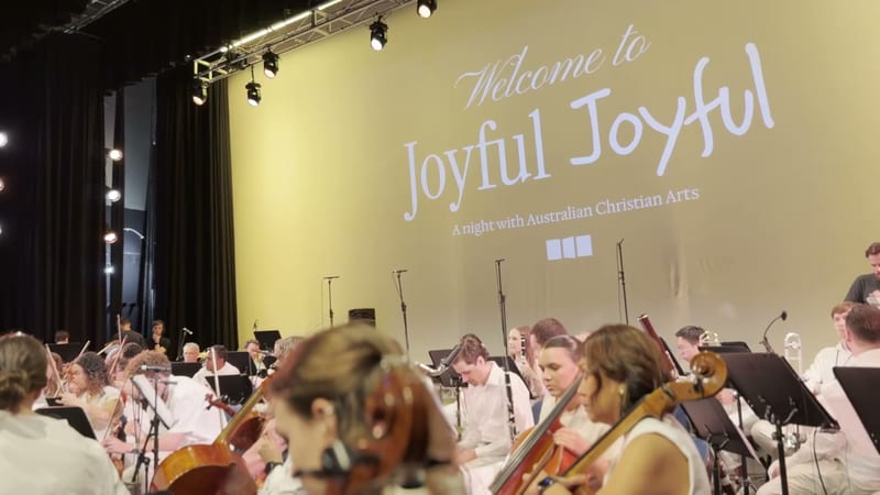 A large ensemble of string players performs during 'Joyful Joyful,' a night with Australian Christian Arts, with the stage backdrop and lighting visible behind them.