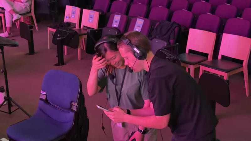 Two people wearing headphones collaborate at a workstation in a concert venue with empty seats.