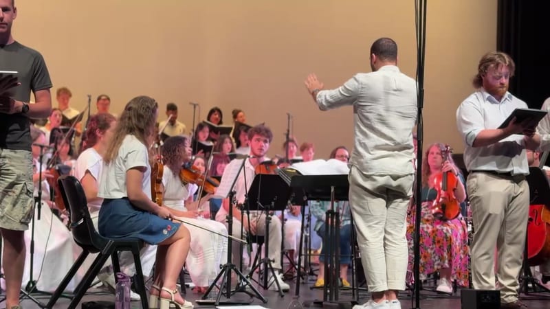 A conductor leads a youth orchestra and choir ensemble during a rehearsal or performance in an auditorium.