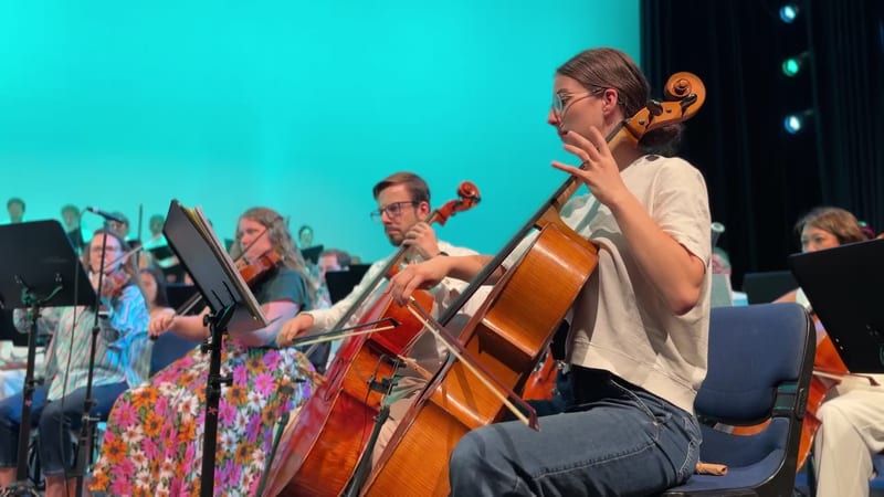 A young cellist performs with the orchestra during a concert against a vibrant turquoise backdrop.