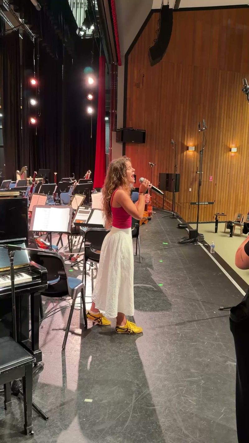 A young female soloist performs at the microphone during an orchestral rehearsal or concert in a professional venue.