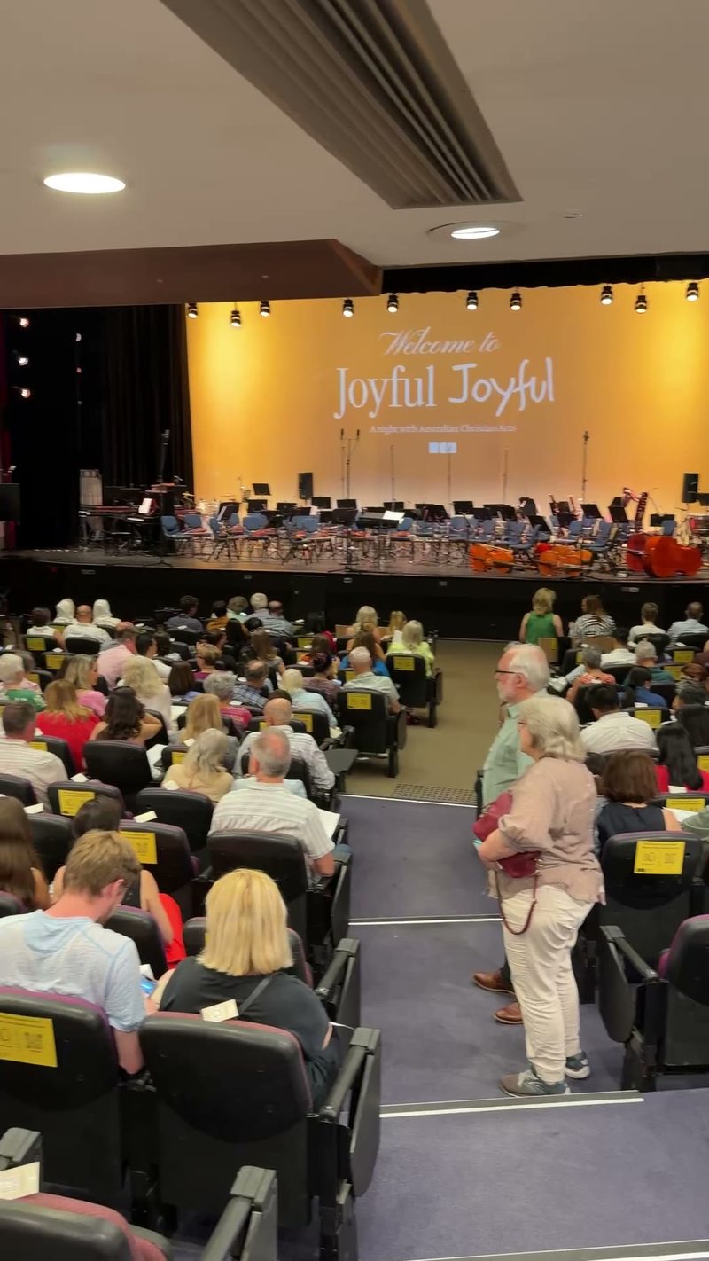 A full auditorium of audience members watches a welcome performance on stage during the Joyful Joyful event.