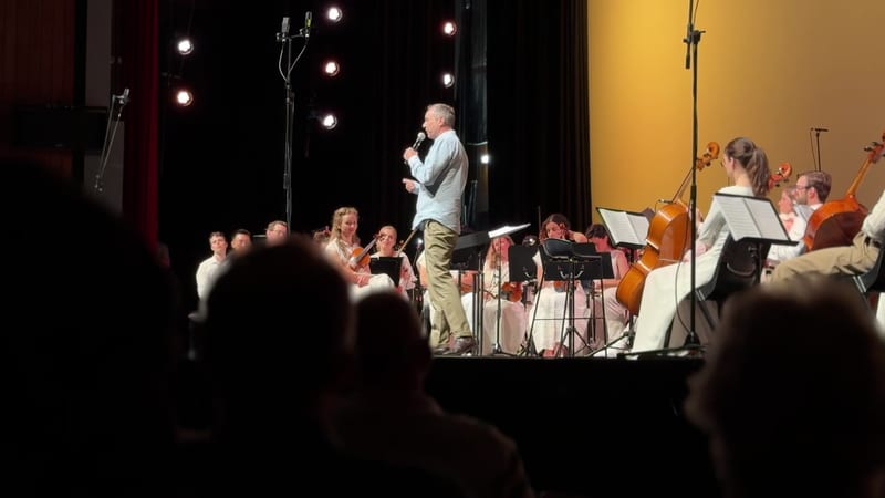 A conductor speaks to the orchestra during a rehearsal or performance with string players visible in the ensemble.