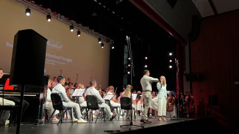 A conductor and soprano soloist perform together with the orchestra during a concert featuring a projection screen backdrop.