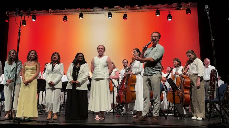 Orchestra and choir members take a bow together on stage after a performance, with the conductor addressing the audience.