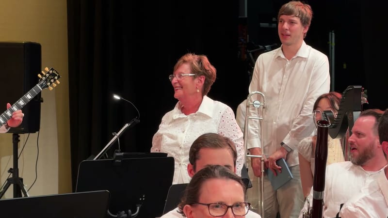 A group of musicians in white shirts perform together on stage with various instruments including guitar, keyboard, trombone, and bass.