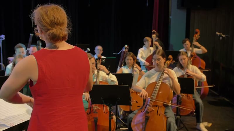 A conductor in a red dress leads a cello section during an orchestral rehearsal or performance.