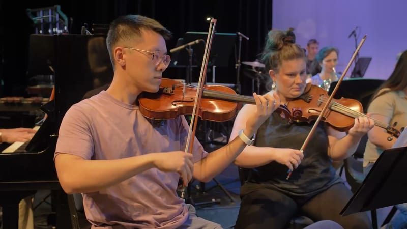 Young violinist in a light purple shirt performs with other string players during an orchestral rehearsal or performance.