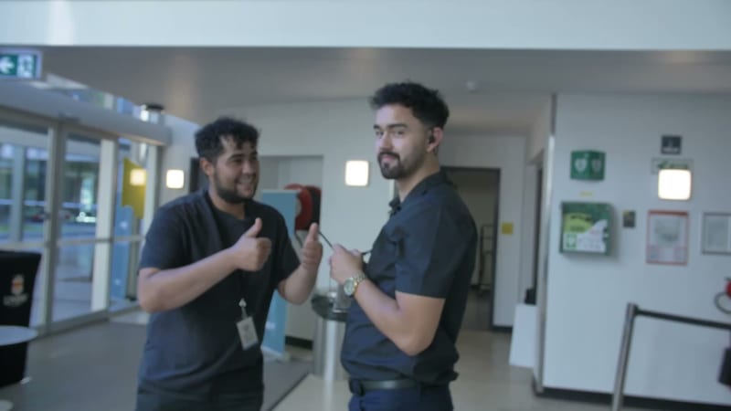 Two men in dark shirts have a conversation in a modern institutional hallway with emergency signage visible.